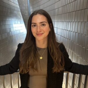 Woman with long brown hair standing and smiling in front of a tiled metallic wall background, representing innovation in occupational health with AI.