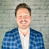 Smiling man in a blue plaid blazer and white shirt standing in front of a light brick wall, representing innovations in occupational health with AI.